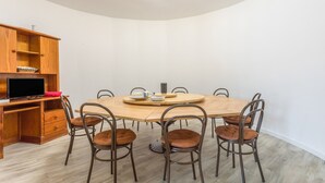 Un coin repas avec une table ronde en bois, six chaises, un téléviseur posé sur un meuble en bois et un mur blanc.