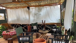 Uma área externa com assentos de madeira, um vaso de terracota grande e um espaço coberto com toldo branco.