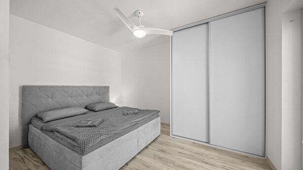 A modern bedroom with a grey upholstered bed, a ceiling fan, and a sliding closet door. - 2 room Apartment with balcony-Nálepkova (Liptovský Mikuláš)