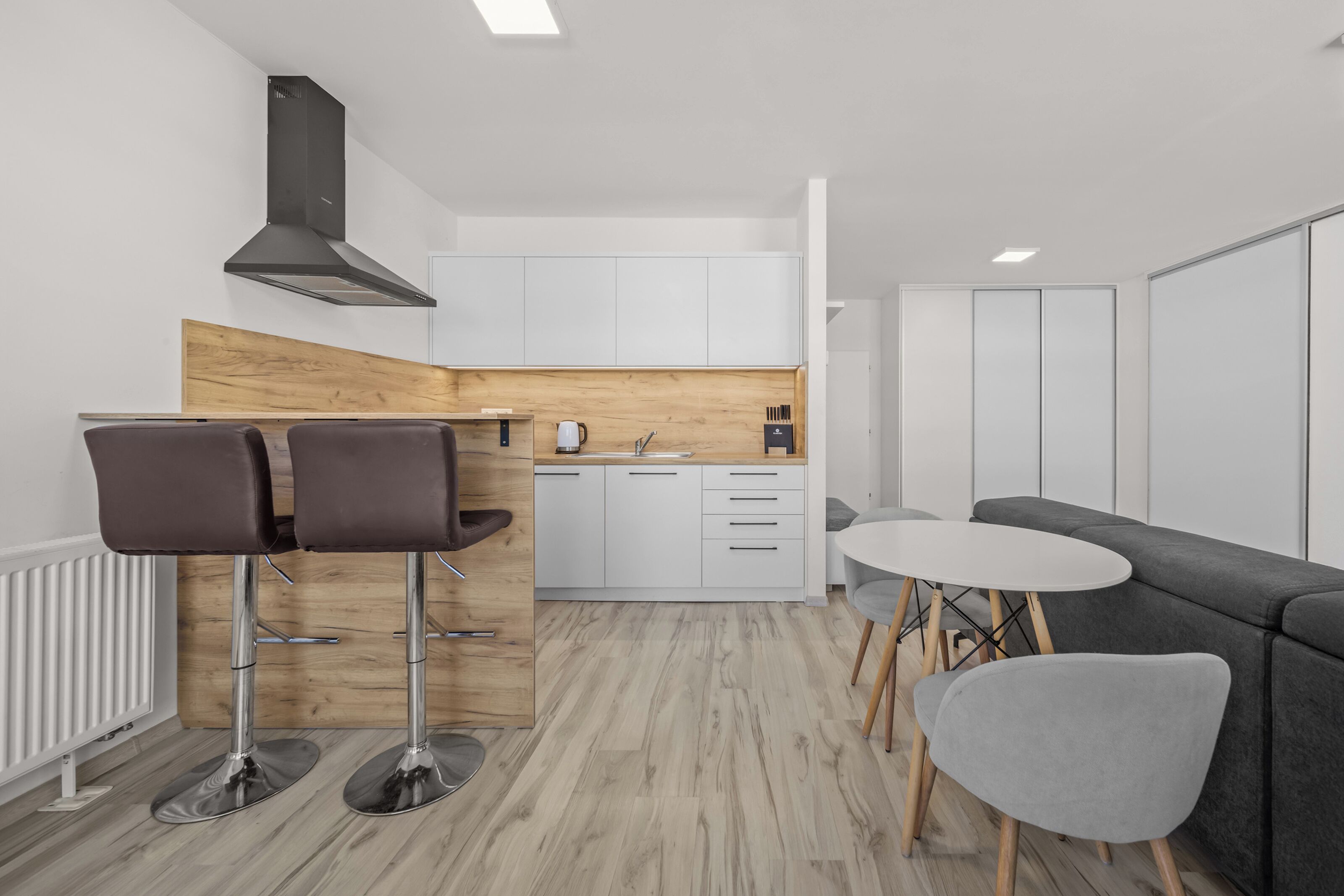 A modern kitchen with white cabinets, a wooden accent wall, a black range hood, and a dining area with a grey sofa and two chairs.