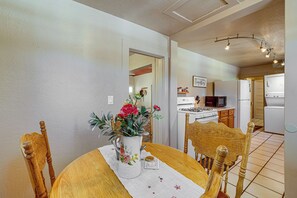 Uma área de refeições com uma mesa de madeira, cadeiras e um vaso com flores. Dá para ver uma cozinha ao fundo, com eletrodomésticos e um letreiro que diz 'Gather Together'.