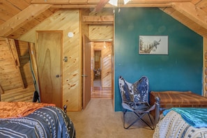Ein Zimmer mit Holzwänden und einer schrägen Decke, einem Bett mit gemusterten Betttextilien, einem Sessel und einem gerahmten Bild an der Wand.