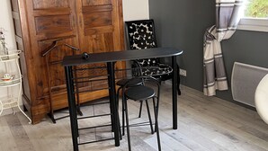 Una habitación con un armario de madera, una mesa metálica negra con dos sillas, un sillón con estampado blanco y negro, y una ventana con cortinas.