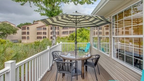 A covered outdoor patio with a table and chairs, a striped umbrella, and a view of a multi-story building in the background. - Ocean-view Retreat Near Barefoot Landing & Beach! (Myrtle Beach)