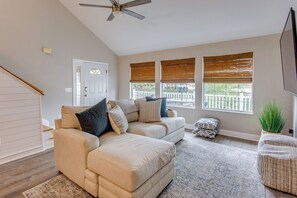 Ein Wohnzimmer mit einem beigefarbenen Ecksofa, einem Deckenventilator und einem großen Fenster mit Jalousien.