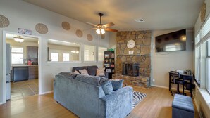 A living room with a grey sofa, a stone fireplace, a flat-screen TV, and a ceiling fan. - Extended East Tulsa Home w/ Fenced Yard (Tulsa)