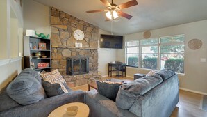 A living room with a stone fireplace, a ceiling fan, and a large window with blinds. - Extended East Tulsa Home w/ Fenced Yard (Tulsa)