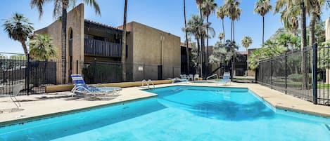 Uma piscina com espreguiçadeiras, cercada por palmeiras e um prédio com varandas.