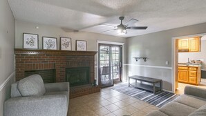 A living room with a fireplace, a ceiling fan, and a kitchen area visible through an open door.