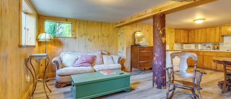 Una sala de estar acogedora con piso de madera, un reposapiés verde, un sofá con cojines, una silla de madera y una cocina con gabinetes de madera y un microondas.