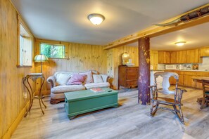 A cozy living room with a wooden floor, a green ottoman, a sofa with cushions, a wooden chair, and a kitchen area with wooden cabinets and a microwave. - Yuba River Retreat for 2! Near Swimming Holes (Washington)