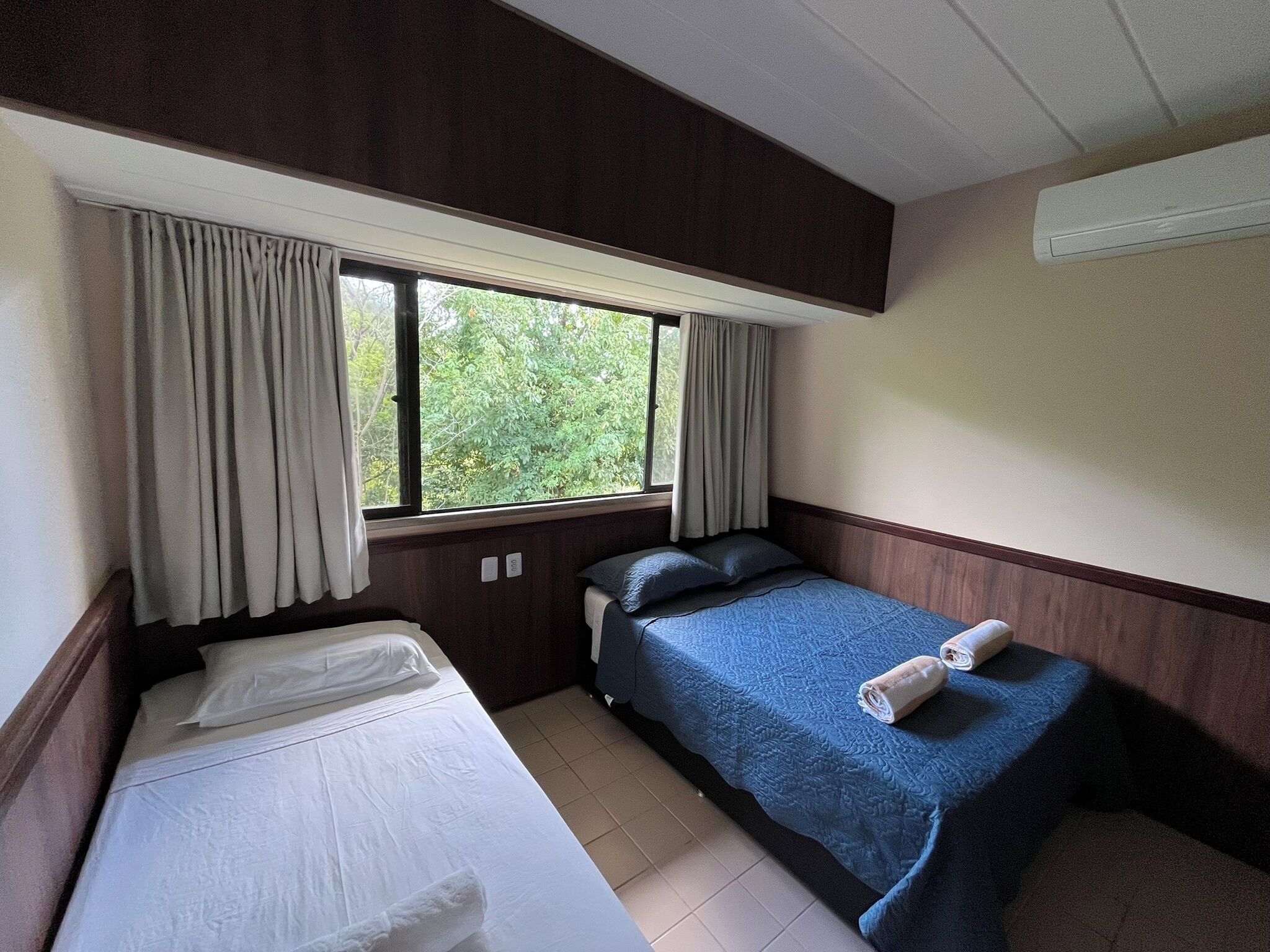 Quarto de hotel com duas camas, uma janela com cortinas e um ar condicionado fixado na parede.