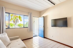 A hotel room with a bed, a TV mounted on the wall, a window with curtains, and a view of palm trees outside. - Guarajuba, 1-bedroom apartment near the beach (Camaçari)