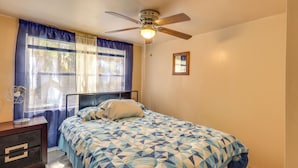 Quarto com cama, ventilador de teto, janela com cortinas, mesa de cabeceira com abajur e um quadro pendurado na parede.