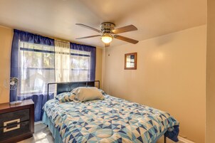 Un dormitorio con cama, ventilador de techo, ventana con cortinas, mesita de noche con lámpara y un cuadro en la pared.