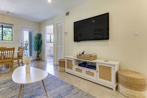 Ein Wohnzimmer mit einem an der Wand befestigten Flachbildfernseher, einer weißen Unterhaltungseinheit mit elektronischen Geräten, einem Essbereich mit Holzmöbeln und einer großen Topfpflanze.