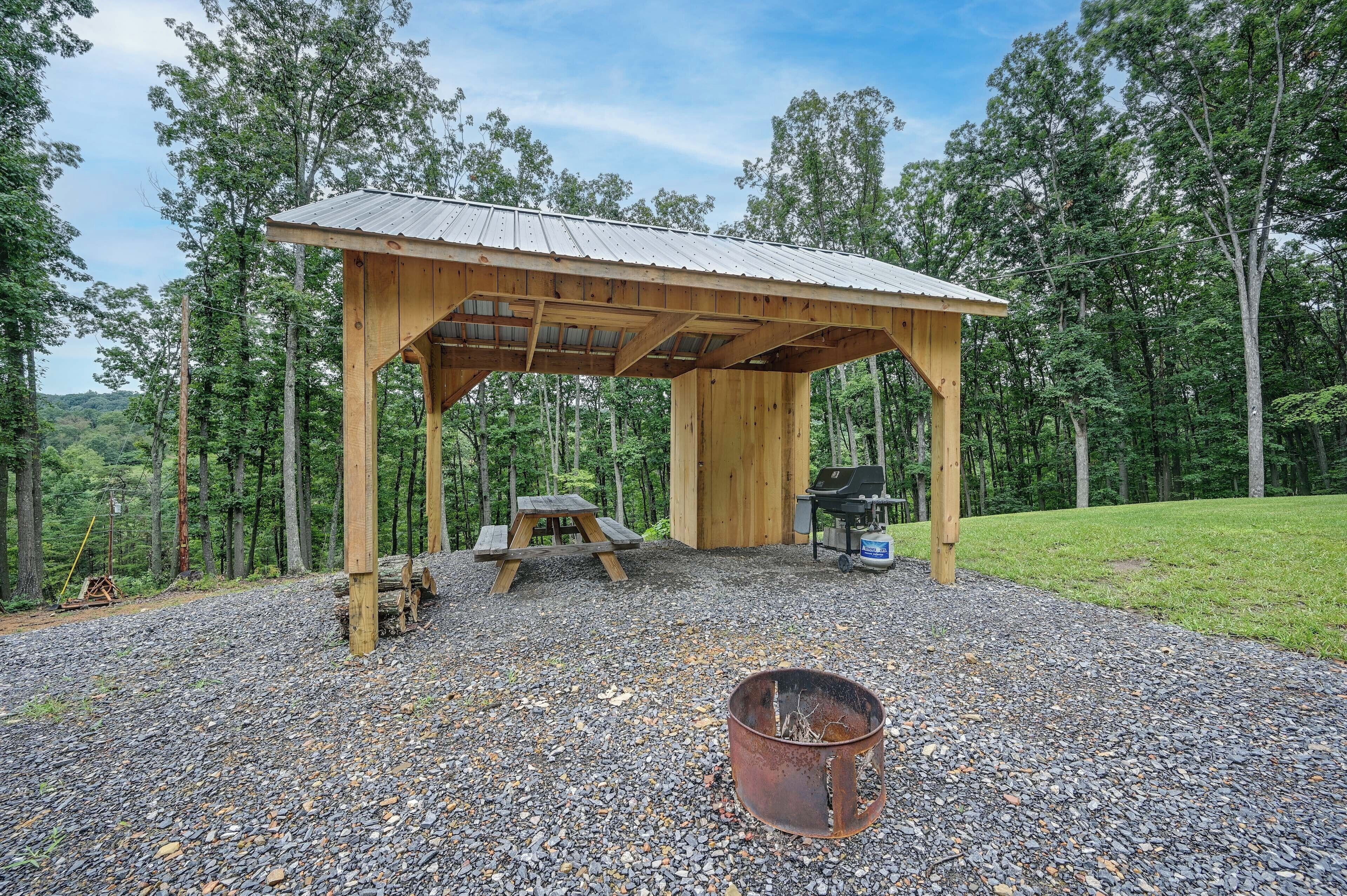 A wooden pavilion with a metal roof, a picnic table, and a grill, surrounded by a gravel area and trees.