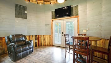 A room with a leather sofa, wooden dining table, chairs, a television mounted on the wall, and a wooden ceiling.