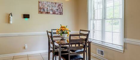Un coin repas comprenant une table, des chaises, un vase de fleurs, un extincteur fixé au mur et une fenêtre avec des stores.