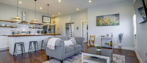 A modern living room with a grey sofa, coffee table, and a kitchen area with a white backsplash and stainless steel appliances.