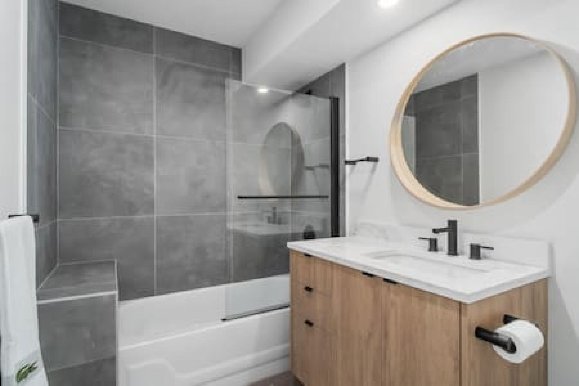 Une salle de bain moderne avec une douche à parois de verre, un meuble-lavabo en bois doté d’un plan de travail blanc et un miroir rond.