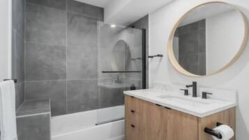 Une salle de bain moderne avec une douche à parois de verre, un meuble-lavabo en bois doté d’un plan de travail blanc et un miroir rond.