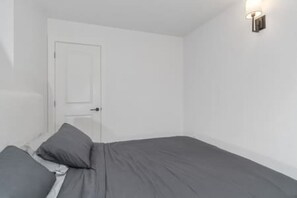 Een eenpersoonsbed met een grijze matras en kussens, een witte deur en een wandlamp.