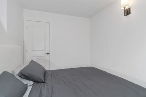 A single bed with a grey mattress and pillows, a white door, and a wall-mounted light fixture.
