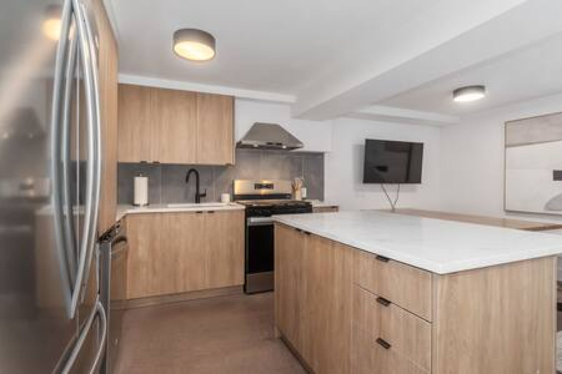 A modern kitchen with wooden cabinets, a stainless steel refrigerator, a built-in oven, and a flat-screen TV mounted on the wall.