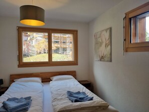 Un lit double avec du linge de lit blanc, une tête de lit en bois et une fenêtre donnant sur un immeuble et des arbres.