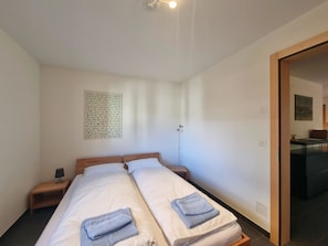 A double bed with two pillows and a blue towel, a bedside table with a lamp, a wall-mounted artwork, and a door leading to a bathroom. - Pastis Appartement/Fewo Appartement/Fewo by Interhome (Saas-Fee)
