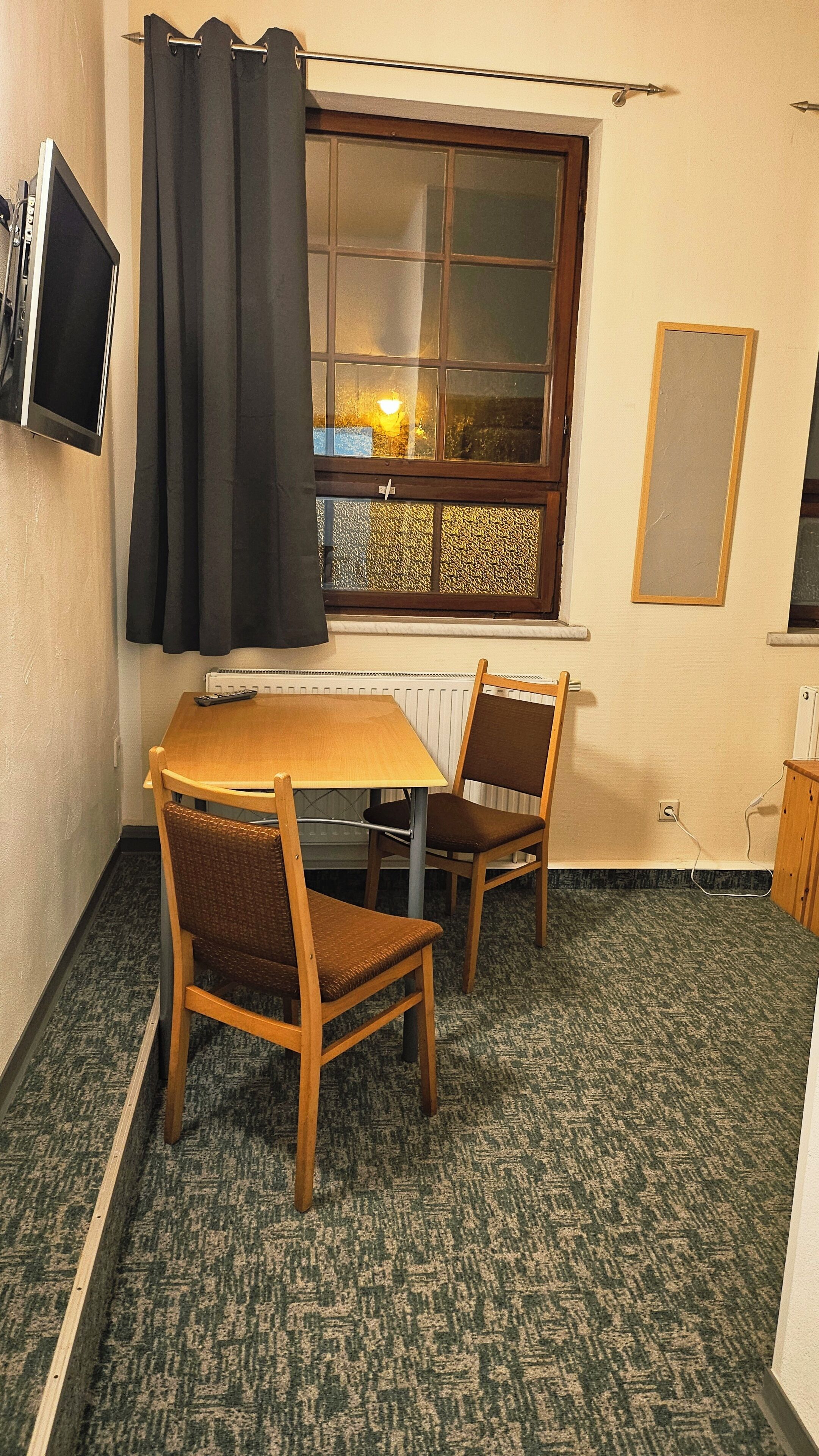 A small room with a wooden table and two chairs, a window with curtains, a radiator, and a television mounted on the wall.