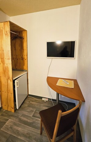 A small room with a wooden desk, a chair, a television mounted on the wall, and a wooden wardrobe.