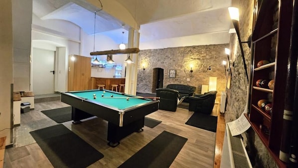 Una mesa de billar en una sala con pared de piedra, zona de bar y área de estar con chimenea.