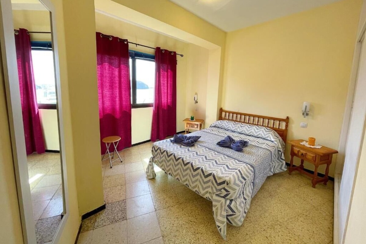 A bedroom with a bed, a small table, a chair, and a window with curtains.