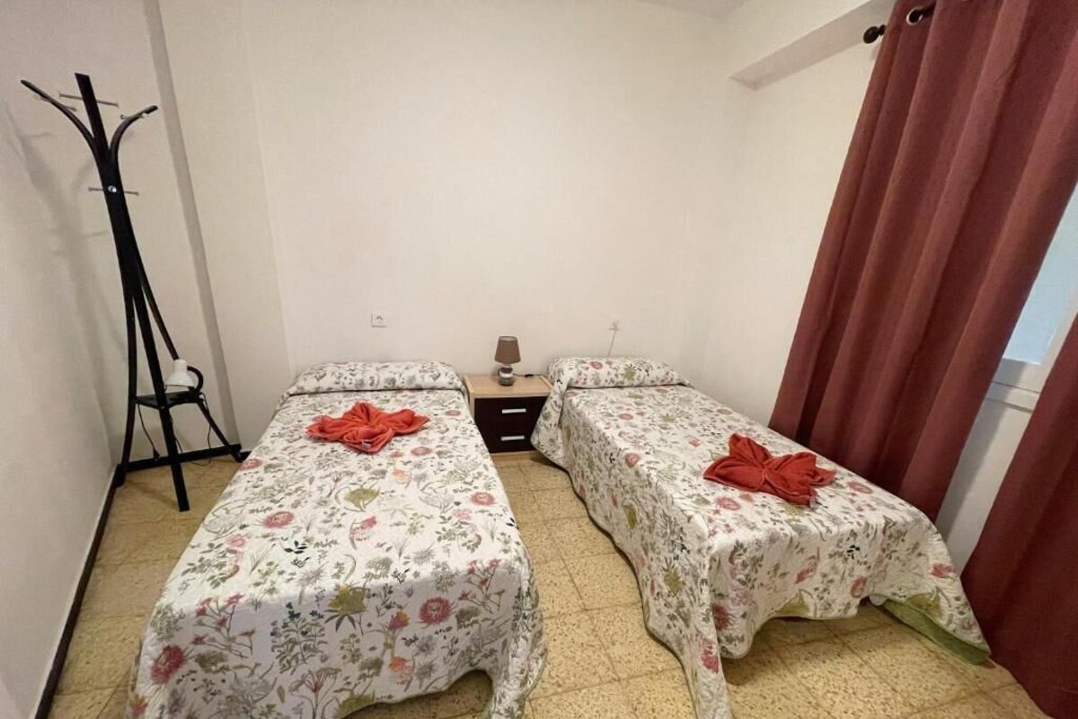A small room with two single beds, a coat rack, a nightstand with a lamp, and a window with curtains.