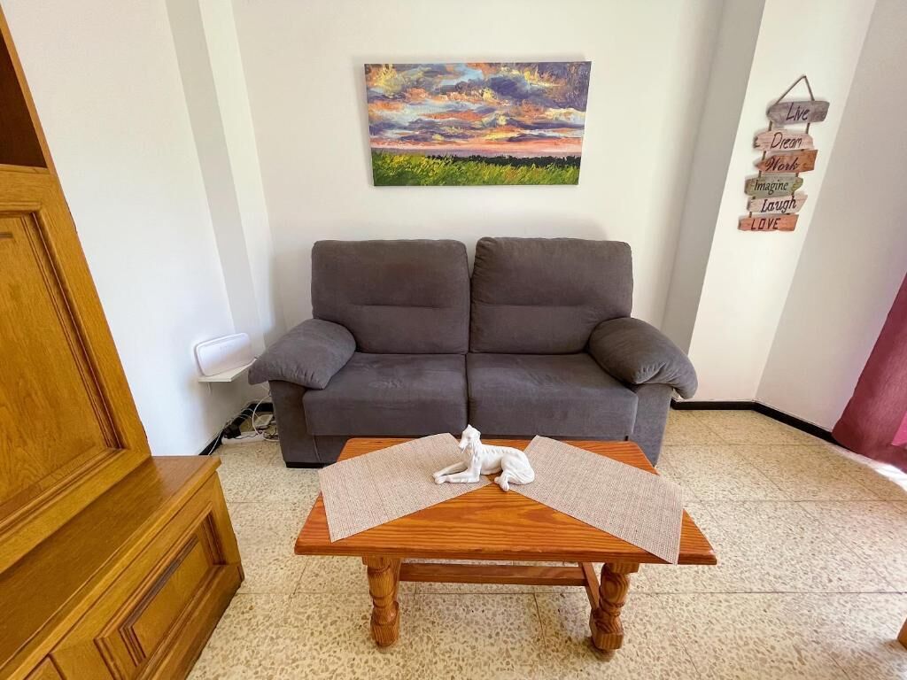 A living room with a grey sofa, a wooden coffee table with a white ceramic dog, a painting of a sunset, and a wall with motivational words.