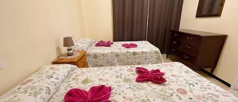 A hotel room with two beds, each with a floral bedspread and a pink towel. There is a wooden nightstand with a lamp, a dresser, and a mirror on the wall.