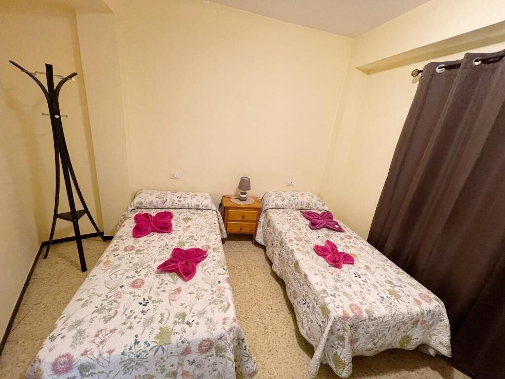 Una habitación pequeña con dos camas, una mesita de noche, una lámpara y un perchero.