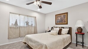 A bedroom with a large bed, a ceiling fan, a window with curtains, a bedside table with a lamp, and a framed picture on the wall.