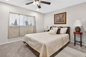 A bedroom with a large bed, a ceiling fan, a window with curtains, a bedside table with a lamp, and a framed picture on the wall.