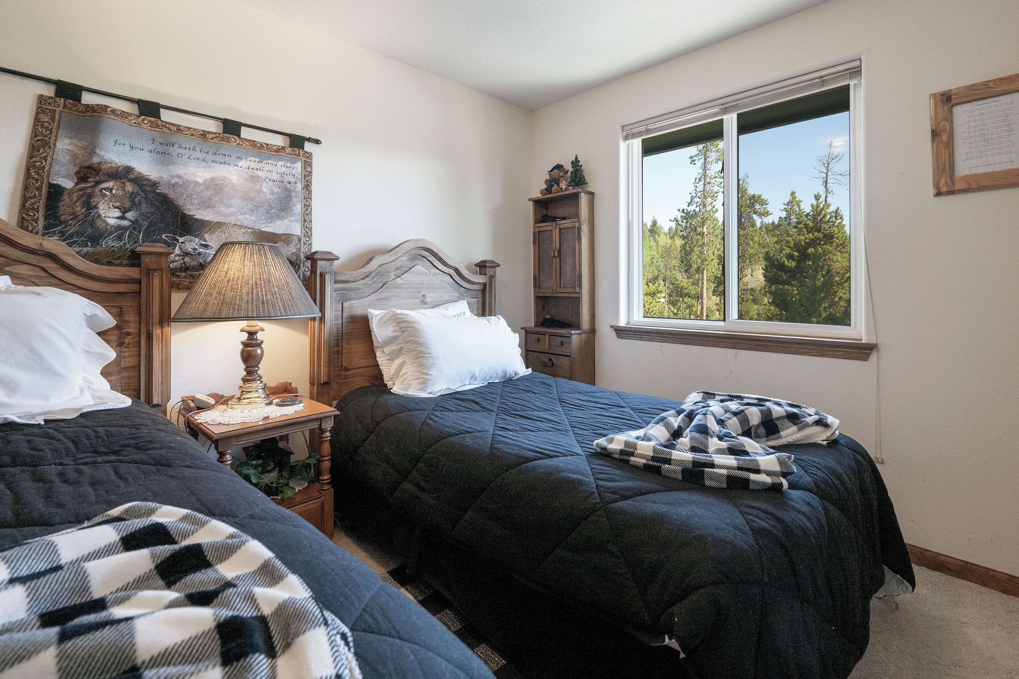 A bedroom with two beds, a nightstand with a lamp, a wooden headboard, a window with a view of trees, and a wall-mounted tapestry.