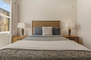 A neatly made bed with a patterned bedspread and pillows, flanked by two bedside tables with lamps, in a room with a wooden headboard and a window with curtains. - Adventure Loft: Pool Views! Games! (Hurricane)