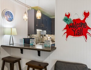 A kitchen with a clock, bar stools, a welcome sign, and a wall-mounted light fixture. - Gulf Village #203 | Gulf Shores Condo with + Community Pool (Gulf Shores)
