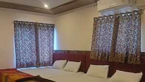 Una habitación de hotel con una cama grande, dos ventanas con cortinas estampadas, un ventilador de techo y un aire acondicionado.
