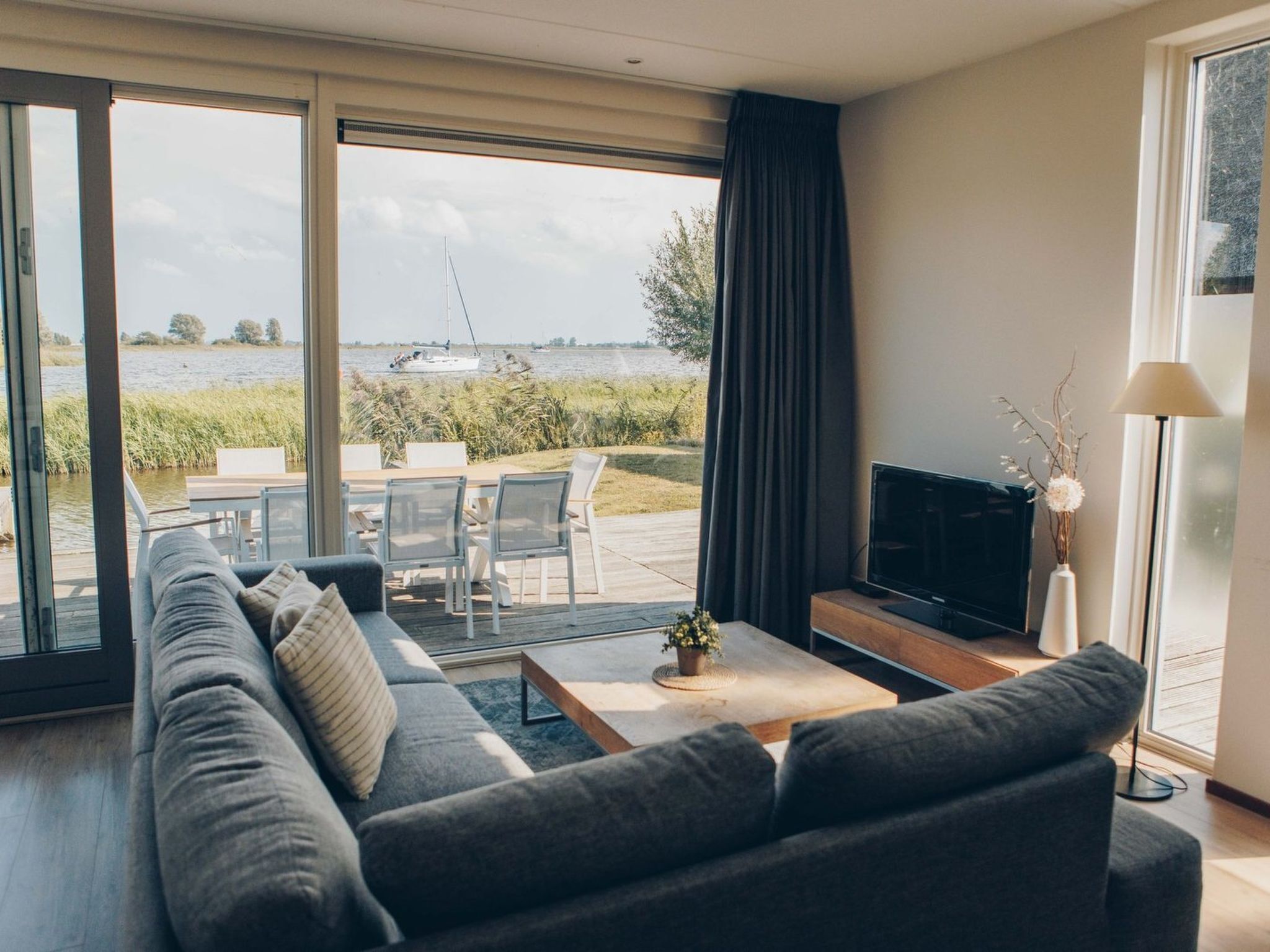 Una sala de estar moderna con un sofá grande, un televisor de pantalla plana y vistas a un lago con un velero.