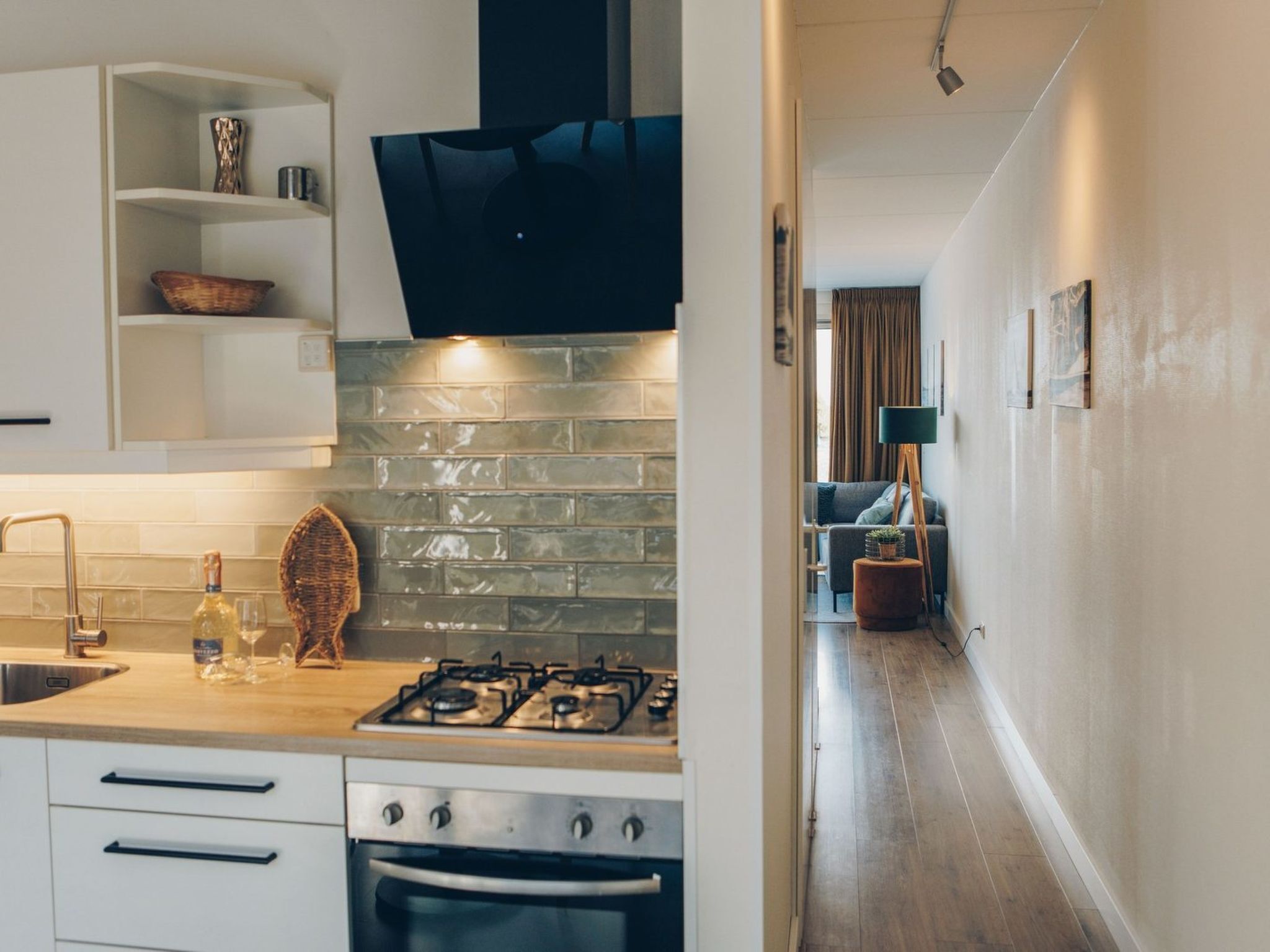 Cocina moderna con estufa a gas, horno y muebles blancos. Se aprecia un protector contra salpicaduras de piedra y una campana extractora negra. En el fondo, se ve un rincón comedor con un sofá y una lámpara.