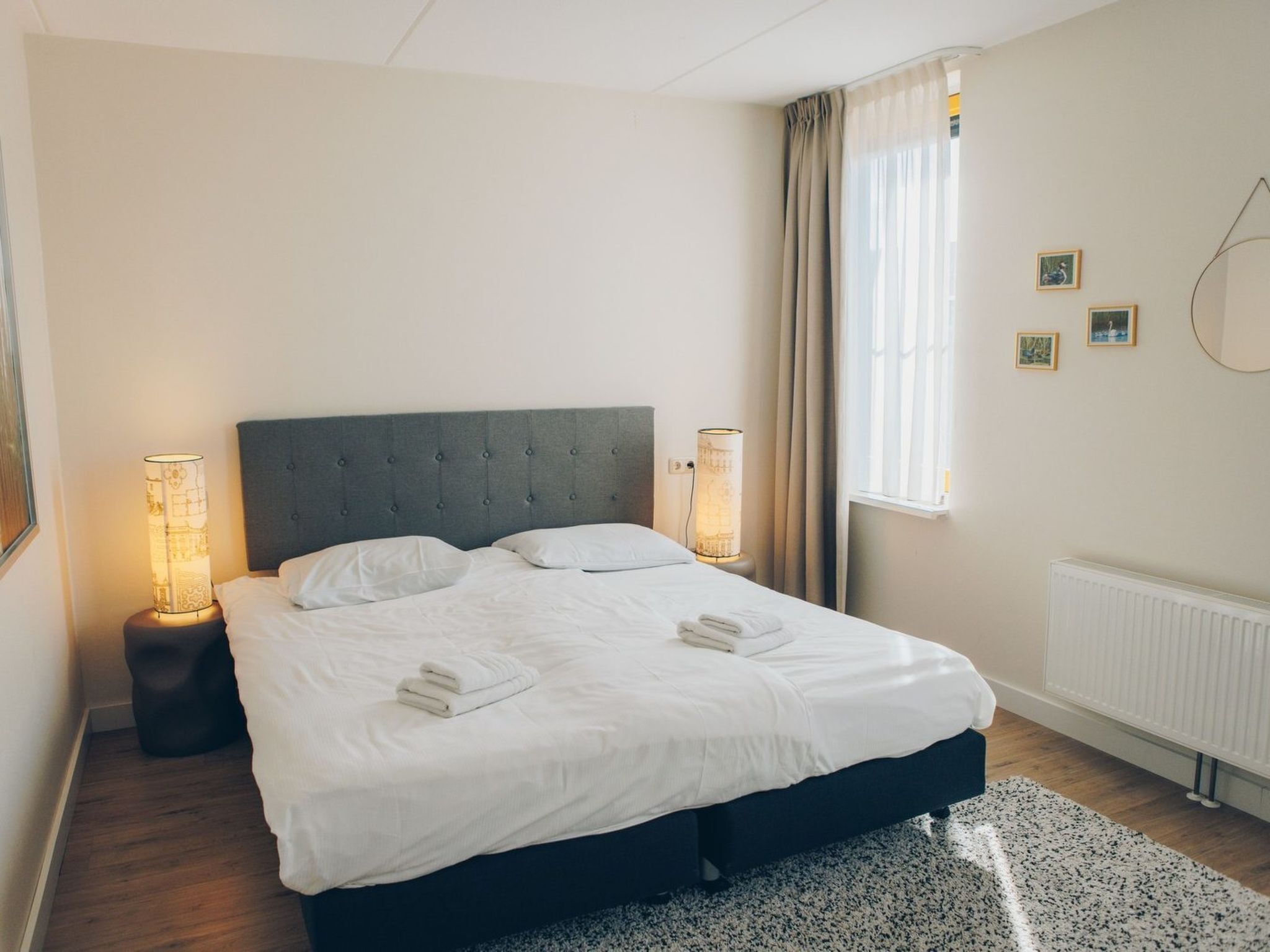 Una cama bien hecha con sábanas blancas y dos almohadas, un cabecero tapizado en gris, una lámpara de noche y un radiador en la pared.