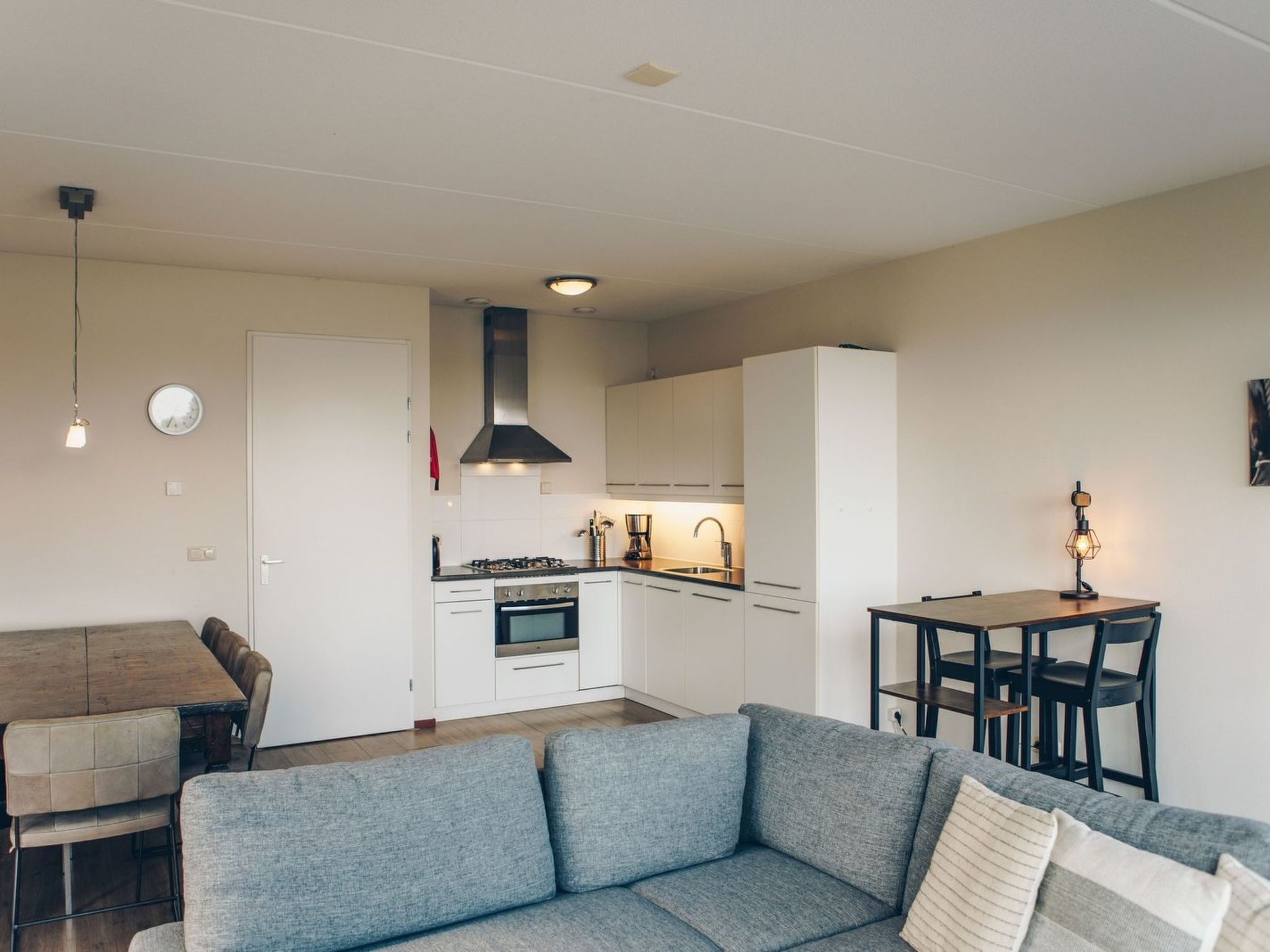 Una cocina moderna con armarios blancos, una estufa y un fregadero. Hay un comedor con una mesa y sillas de madera. Un sofá gris está en primer plano.