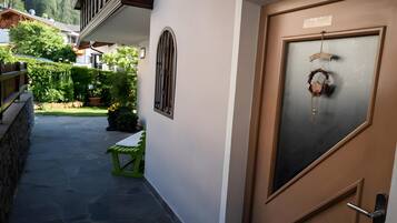Ein Korridor mit einer grünen Bank, einer mit einem Kranz geschmückten Tür sowie Blick auf einen Garten und Gebäude im Hintergrund.
